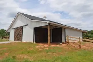 Exterior view of a custom horse barn built by Cox Builders, featuring white vertical siding, dark wooden sliding doors, and a black shingle roof with a cupola. The barn includes an open sheltered bay and surrounding fencing, designed for both function and durability.