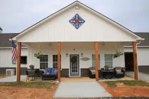 Welcoming custom home by Cox Builders with a modern farmhouse design. The exterior features crisp white board-and-batten siding accented by stonework at the base and warm wooden posts supporting the front porch. Comfortable outdoor seating areas with hanging plants create an inviting atmosphere, while a patriotic American flag adds a touch of charm. A decorative geometric barn quilt detail above the entry enhances the home’s character, making this build a perfect blend of style, comfort, and personality.